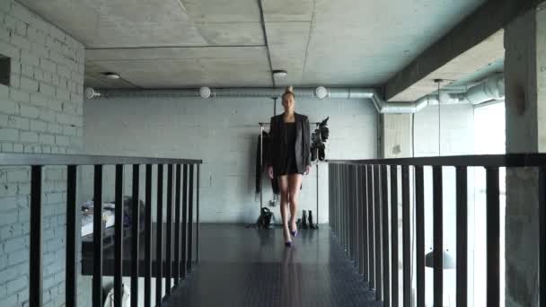Kadın model, evde ayakkabısıyla karanlık bir podyum boyunca yürüyor ve bir kıyafet gösteriyor. Kadın sarışın ve saçı topuz şeklinde toplanmış. Uzun boylu ve incedir. Çatı katı biçimi — Stok video