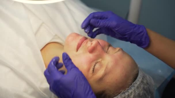 Kosmetolog tillämpar transparent komposition på kvinnor ansikte med cirkulära rörelser innan förfaranden. Rengöring av orenheter. Lilac handskar är på läkarnas händer. Vit, ren hud — Stockvideo