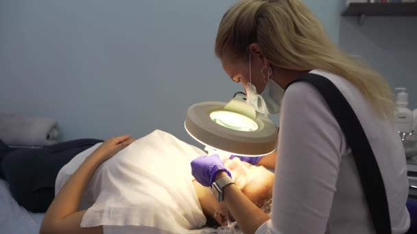 Cosmetólogo realiza el procedimiento para la mujer. El médico rubio mira a través de la lupa y realiza el procedimiento de limpieza de la piel. Examina la piel bajo lupa. Tratamiento de la piel — Vídeo de stock