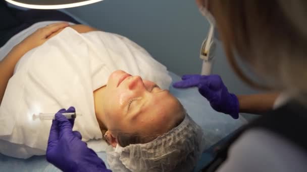 Dokter brengt helende stof voor acne van injectiespuit met gladmakende bewegingen op een eerlijk gezicht. Cosmetoloog heeft masker aangebracht en doet gezichtsmassage. Lamp boven de patiënten gezicht. Acne behandeling — Stockvideo