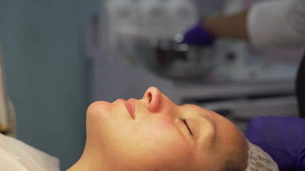 Cosmetologist traite les femmes visage avec des éponges blanches. Nettoyer la peau des impuretés. Rendez-vous chez le médecin. Cap est mis sur ses cheveux, gants lilas sont sur les mains des médecins. Peau blanche et propre — Video