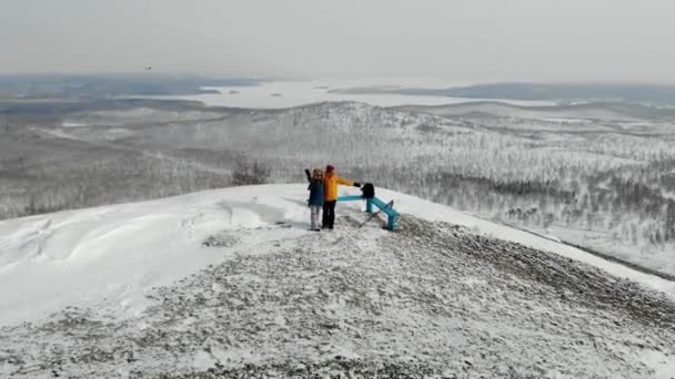 Mężczyzna i kobieta stoją na szczycie pokrytego śniegiem wzgórza. Zatrzymali się, by odpocząć. Machają rękami. Widać stoki śnieżne, drzewa i kominy fabryczne. Dym wydobywa się z rur. Widok z lotu ptaka — Wideo stockowe