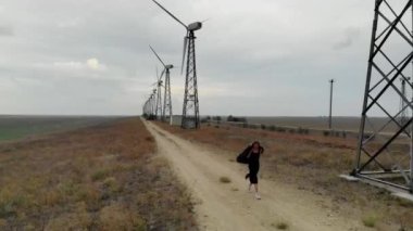 Rüzgar türbinlerinin hava görüntüsü. Boş tarlanın arka planında çiftlik rüzgârı türbinleri. Kadın onlarla yürüyor. Sakin hava ve rüzgar türbinleri hareket olmadan çalışmaz. Mavi gökyüzünde bulutlar var