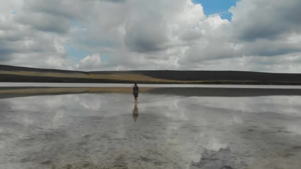 Widok z powietrza na beżowe słone jezioro. Kobieta w sukience chodzi po wodzie jak po lustrze. Woda jeziora odzwierciedla błękitne niebo i unoszące się chmury. Wyjątkowa natura. Opuszczony — Wideo stockowe