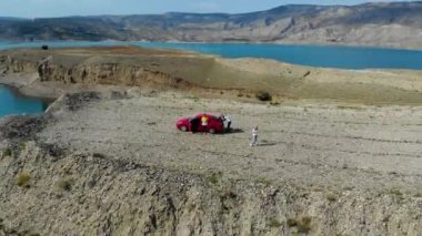 Baraj gölünün havadan görünüşü. Gök mavisi suyu olan yapay göl. Höyükler adalar gibidir. Karada kırmızı bir araba var. Bulutlar suya yansıyor. Dağlar uzaktan görülebilir.. 