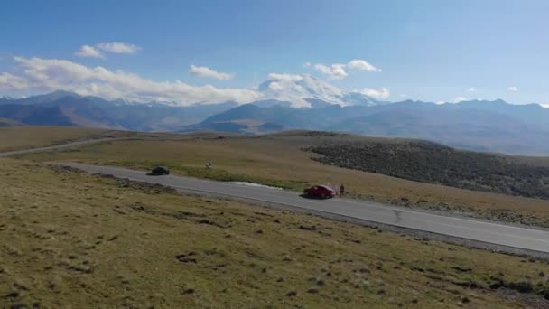 Widok z lotu ptaka na drogę biegnącą przez łańcuch górski Kaukazu, region Elbrus. Samochody stoją na drodze wśród zielonych łąk. Słońce oświetla szczyty gór. — Wideo stockowe