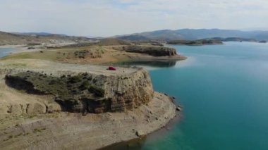 Baraj gölünün havadan görünüşü. Gök mavisi suyu olan yapay göl. Höyükler adalar gibidir. Karada kırmızı bir araba var. Bulutlar suya yansıyor. Dağlar uzaktan görülebilir.. 