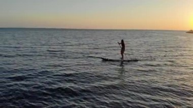 Kadın SUP tahtasının üzerinde yüzüyor. Bikinili bronzlaşmış kadın gemide kürekle kürek çekiyor. Küreği sıkıca elinde tutuyor. Güneşli gün batımı. Deniz gök mavisi renginde. Sıcak hava