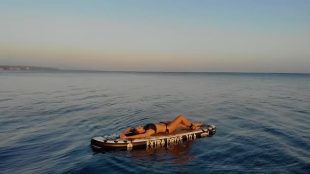 Kvinnan flyter på SUP-brädet. Vacker solbränd kvinna ligger på rygg ombord i bikini och simmar smidigt på små vågor. En åra ligger bredvid den. Havet är azurfärgat. Varmt väder. Sommar — Stockvideo