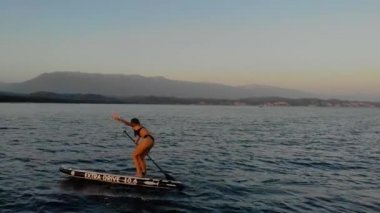 Kadın SUP tahtasının üzerinde yüzüyor. Bikini giymiş, kürek çekiyor, sendeliyor ve suya düşüyor. Küreği sıkıca elinde tutuyor. Deniz gök mavisi rengidir. Sıcak hava
