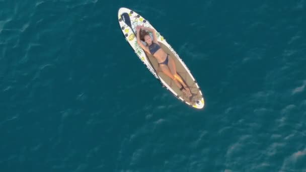 Frau schwimmt auf SUP-Board. Schöne, braun gebrannte Frau liegt im Bikini auf dem Rücken an Bord und schwimmt geschmeidig auf kleinen Wellen. Daneben liegt ein Ruder. Das Meer ist azurblau. Warmes Wetter. Sommer — Stockvideo