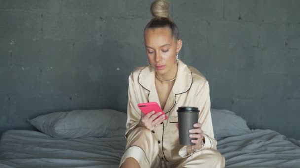 Młoda kobieta pije kawę i trzyma telefon. Dziewczyna w pięknej jedwabnej piżamie pije kawę lub herbatę z tekturowego kubka. Ma smartfona w rękach. Dzień dobry pani. — Wideo stockowe