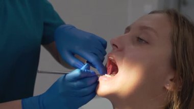 Ortodontist, aletle diş tellerinden pembe elastik bant çıkarıyor. Doktorların elleri lastik eldivenin içinde. Hastaların ağzı sonuna kadar açık. Kadın sonuçtan memnun ve geniş bir şekilde gülümsüyor. Parantezler
