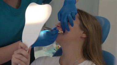 Ortodontist hastaların diş tellerine kauçuk çubuklar takar. Kadın aynayı gözünün önünde tutuyor ve doktorun ne yaptığına bakıyor. Doktorların elleri lastik eldivenin içinde. Aletlerin ellerinde. Parantezler
