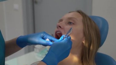 Ortodontist diş tellerindeki elastik bantları aletle tamir eder. Doktorların elleri lastik eldiven giymiş. Hastaların ağzı sonuna kadar açık. Kadın sonuçtan memnun ve geniş bir şekilde gülümsüyor. Parantezler