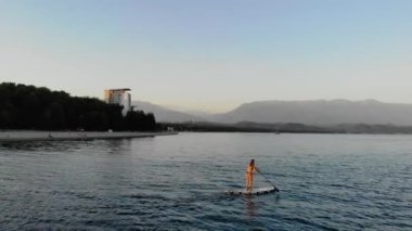 Kadın SUP tahtasının üzerinde yüzüyor. Bikinili bronzlaşmış kadın gemide kürekle kürek çekiyor. Küreği sıkıca elinde tutuyor. Güneşli gün batımı. Deniz gök mavisi renginde. Sıcak hava