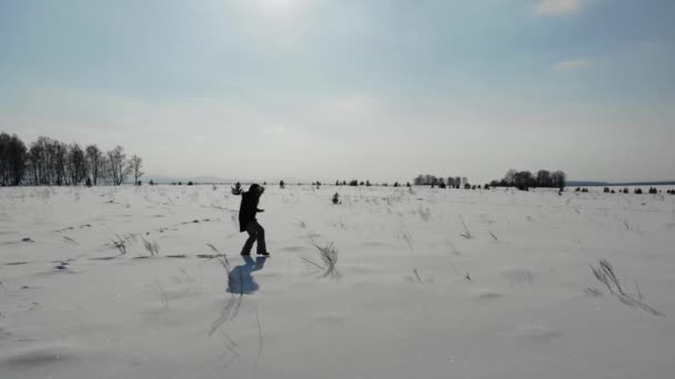 A mulher atravessa o campo de neve. Ela de casaco azul e calças cinza está andando através do campo que está coberto de neve. Ela tem um capuz na cabeça. Seus pés afundam profundamente na neve. É difícil andar. — Vídeo de Stock