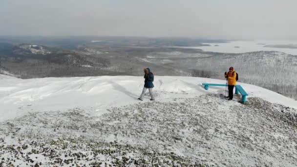 Muž a žena kráčí po pěšině na vrcholu zasněženého kopce. Muž ve žluté bundě chodí s klacky. Nordic walking. V dálce můžete vidět sněhové stromy a jezero. Letecký pohled — Stock video