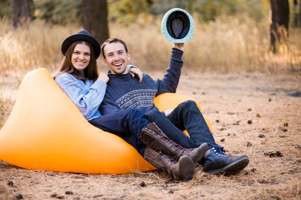 Attractive smiling guy and girl in hat with hands up having fun together, laughing while laying on air sofa Lamzac in autumn forest