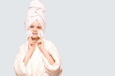 Başında havluyla gülümseyen güzel bir kız çocuğu pamuk bloknotla beyaz arka planda izole edilmiş temizleyiciyle cildi temizliyor. Mutlu kız makyajını sil ve kameraya bak. Sağlıklı, temiz cilt bakımının keyfini çıkar.