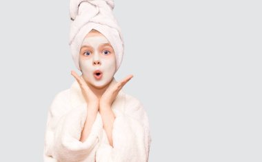 Kafası karışmış, beyaz arka planda izole edilmiş kız çocuğu ellerini yana açıyor, güzel görünmek için yüz maskesi takıyor, beyaz bornoz giyiyor ve kafasına havlu geçiriyor, bir konuda tereddüt ediyor.