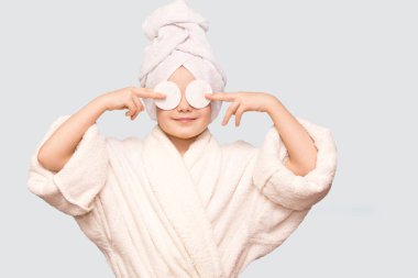 Kafasında havluyla gülümseyen güzel bir kız çocuğu pamuk tutacağı ile cildi temizleyici ile temizliyor. Mutlu kız makyajını temizler sağlıklı cilt bakımının tadını çıkarır.