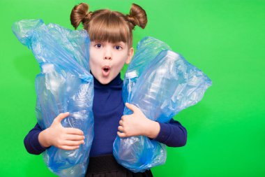 Kafası karışmış, komik suratlı, yeşil arka planda plastik şişelerle dolu çöp torbaları tutan küçük bir çevreci kız.
