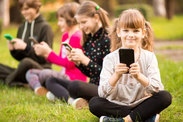 Outdoor portret pozytywnego inspirowane dziecko, dziewczyna siedzi na trawie z nogami skrzyżowane i za pomocą telefonu komórkowego w towarzystwie przyjaciół — Zdjęcie stockowe