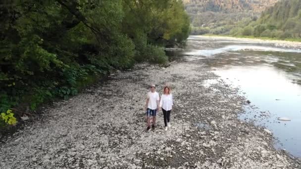 Vue aérienne de drone de jeune couple tenant la main et marchant sur la plage de la rivière de montagnes avec forêt verte autour. — Video