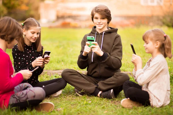 Grupo de niños ocupados mirando sus teléfonos mensajes de texto SMS y jugar juegos sentados con las piernas cruzadas en la hierba fuera en el parque — Foto de Stock