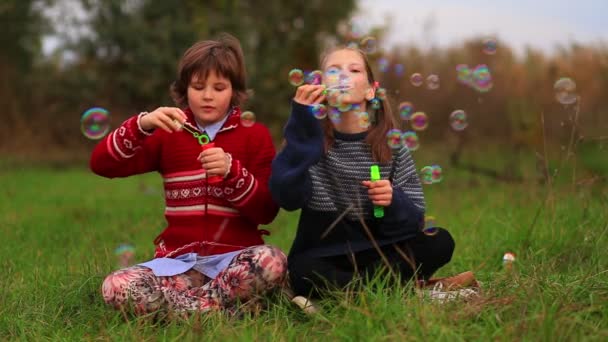 Cute dziecko dziewcząt siedzi na trawie ze skrzyżowanymi nogami i dmuchanie baniek mydlanych w parku. Zabawa bańkami mydlanymi — Wideo stockowe