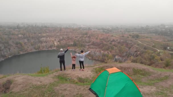 Активная концепция семейного путешествия. Вид с воздуха на молодых родителей с дочерью, идущих рядом с палаткой кемпинга с поднятыми руками во время похода рядом с озером. — стоковое видео