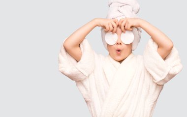 Şaşkın yüzü ve havlusu olan güzel bir kız çocuğu pamuk tutacağı ile cildi temizleyici ile temizliyor. Mutlu kız makyajını temizler sağlıklı cilt bakımının tadını çıkarır.