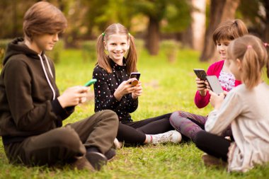 Telefonlarına bakıp mesaj atan ve oyun oynayan bir grup meşgul çocuk parkta çimlerin üzerinde bacak bacak üstüne atarak oturuyor.