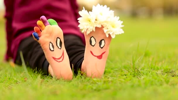 Little child girl sits on grass with painted feet with funny comic faces — Stock Video