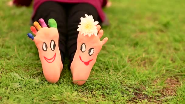 Menina criança senta-se na grama com os pés pintados com caras engraçadas cômicas — Vídeo de Stock