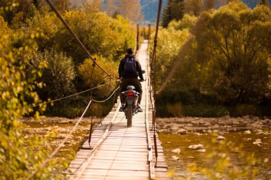 Tanımlanamayan bir adam, asma köprüde, dağların üzerinde motosikletle geziyor.