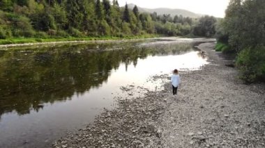 Genç bir kadının hava aracı görüntüsü yaz sabahı dağlarda nehir boyunca yürüyor.
