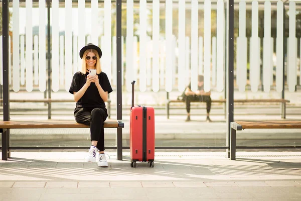 Siyah günlük kıyafetler, güneş gözlüğü ve kırmızı bavullu şapka giyen gezgin turist kadın internet üzerinden bilet almak ve istasyonda oturmak için cep telefonu kullanıyor. Kadın yolcu hafta sonları yurtdışına seyahat ediyor. Hava