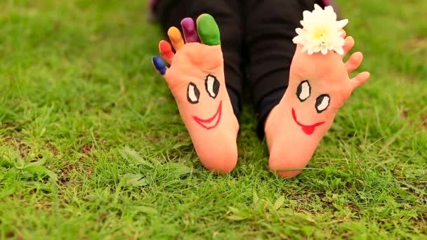 Menina criança senta-se na grama com os pés pintados com caras engraçadas cômicas — Vídeo de Stock