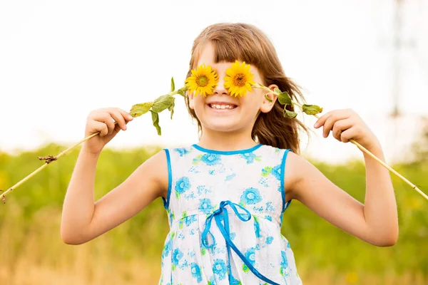 Linda niña usar vestido con girasol en el campo de verano. Feliz niña esconde el ojo con girasol. Concepto vacaciones de verano. —  Fotos de Stock