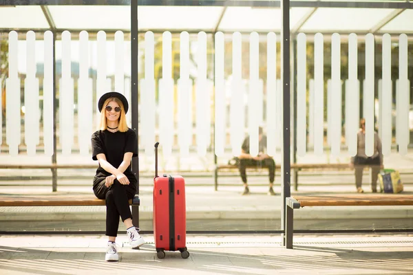 Full length portret van stijlvolle reizende jonge vrouw dragen in totaal zwart met rode koffer wacht vervoer — Stockfoto