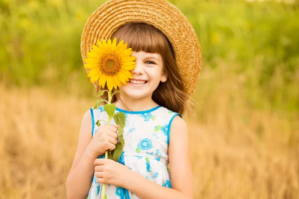 Linda niña usar vestido con girasol en el campo de verano. Feliz niña esconde el ojo con girasol. Concepto vacaciones de verano. —  Fotos de Stock