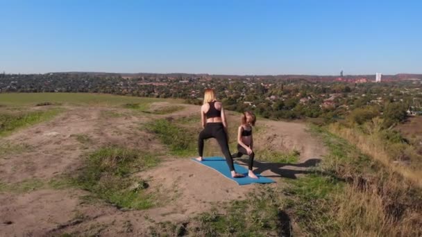 Vue aérienne de drone de mince jeune mère faire des exercices de yoga avec fille enfant sur la haute colline au bord du lac le matin, loin de l'agitation de la ville, donner le bon exemple et inculquer dans ses habitudes saines — Video