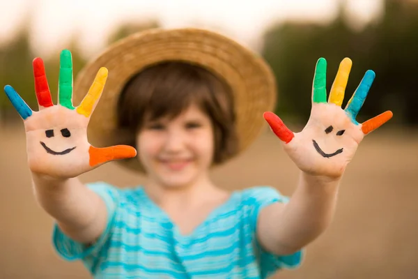 Счастливая улыбчивая маленькая девочка с раскрашенными в смешные цвета руками играет на улице в парке. Сосредоточьтесь на руках — стоковое фото