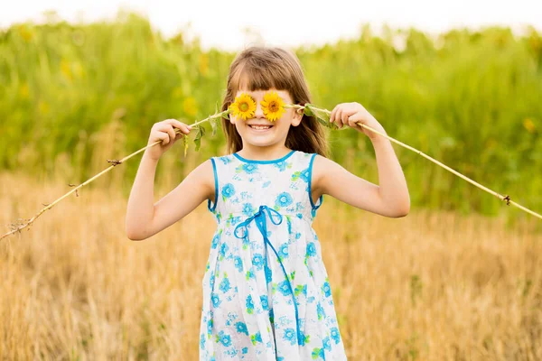 Linda niña usar vestido con girasol en el campo de verano. Feliz niña esconde el ojo con girasol. Concepto vacaciones de verano. —  Fotos de Stock