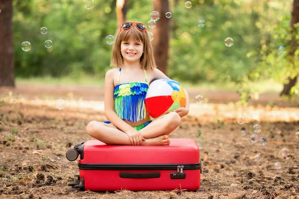 Traveler bambina si siede con le gambe incrociate sulla valigia in occhiali da sole e costume da bagno, pronto a volare al mare e avere vacanze attive — Foto Stock