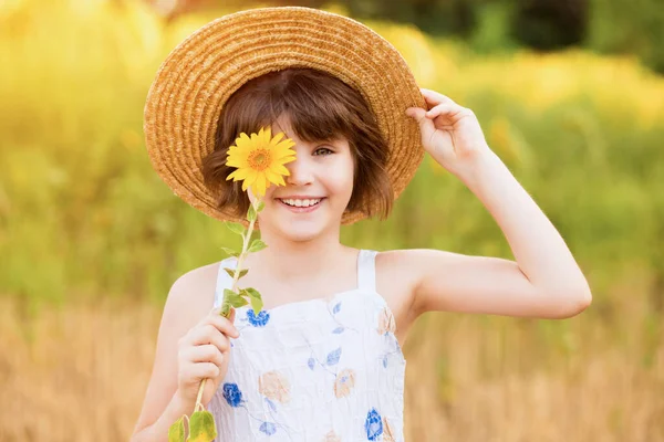 Belle petite fille en chapeau de paille avec des cheveux flottants cachent oeil avec fleur de tournesol, marche en plein air pendant les vacances d'été — Photo