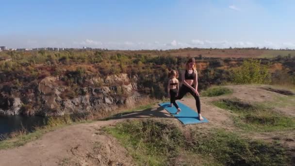 Drohnenbild einer schlanken jungen Mutter, die morgens mit ihrer kleinen Tochter Yoga-Übungen auf einem hohen Hügel am See macht, weit weg von der Hektik der Stadt, mit gutem Beispiel vorangehen und ihre gesunden Gewohnheiten vermitteln kann — Stockvideo