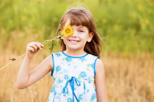 Menina bonito vestir vestido com girassol no campo de verão. Menina feliz esconder o olho com girassol. Férias de verão conceito. — Fotografia de Stock
